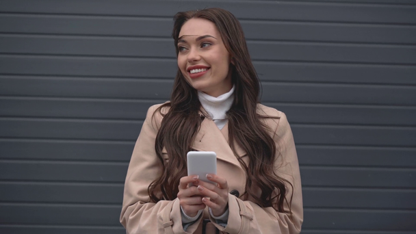 elegante vrouw in beige trenchcoat typen op smartphone bij koude winderige weer lachend - Video