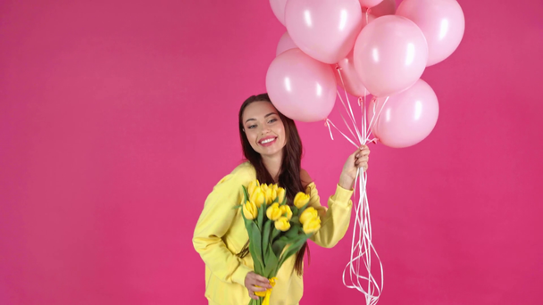 studio shoot of happy young woman holding pink air balloons, smelling yellow tulips and dancing on crimson background - Footage, Video