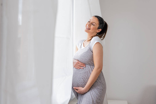 A pregnant woman standing next to the window with hands on her b - Foto, Imagem