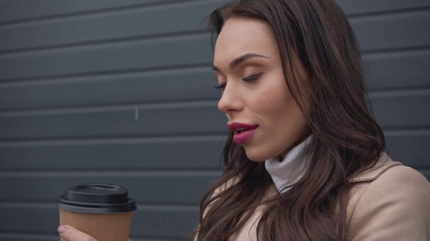 lachende elegante vrouw in beige trenchcoat koffie drinken vanaf papier cup in koude winderige weer - Video