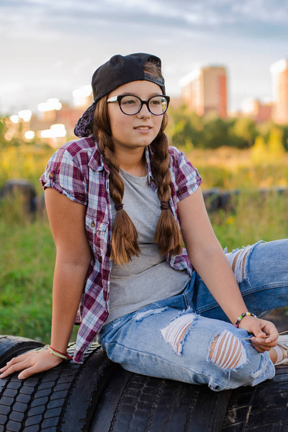 girl teenager in glasses sits the ground city background in the headphones of selfie smartphone - Photo, Image