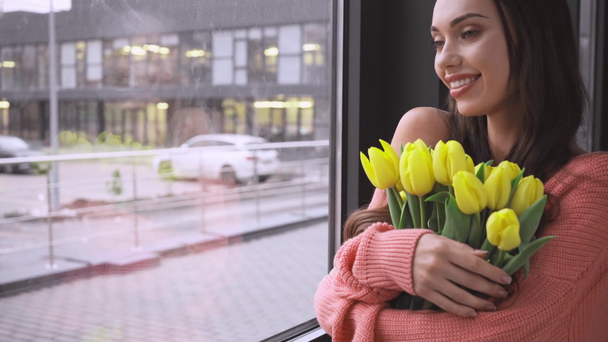 beautiful young woman standing near window and smelling yellow tulips with closed eyes - Filmagem, Vídeo