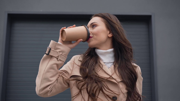 smiling elegant woman in beige trench coat walking and drinking coffee from paper cup in windy weather - Footage, Video