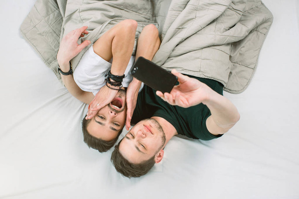 Young gay couple lying under blanket on bed in light room an making selfie on smartphone. Gay couple concept, homosexual - Foto, imagen