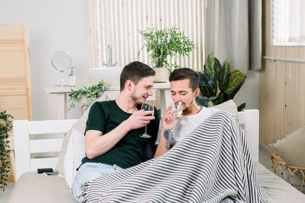 Happy gay couple lying on bed with glasses of wine at home. Gay couple hugging and kissing in bed. Gay Couple Love Home Concept - Foto, Imagen