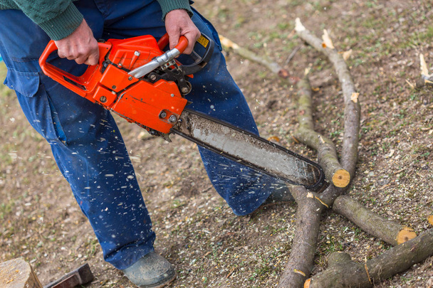 Viejo sosteniendo una motosierra naranja con sus propias manos y cortando una rama colocada en el suelo. motosierra naranja en acción
. - Foto, Imagen