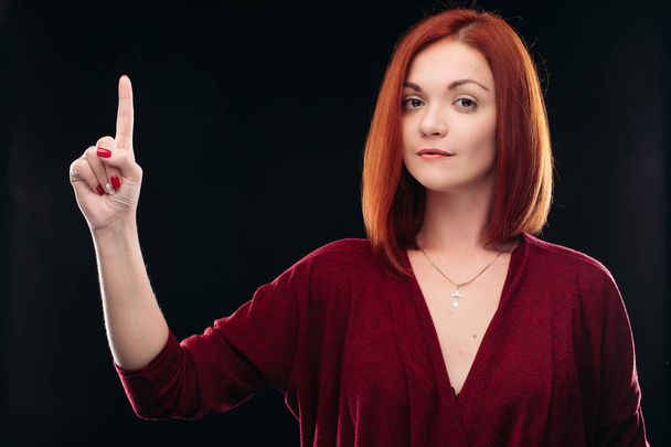 Привлекательная рыжеволосая девушка держит руку с поднятым пальцем
. - Фото, изображение
