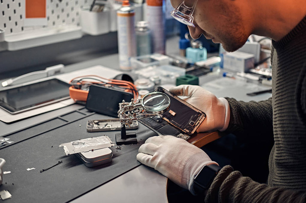 Technician uses a magnifying glass to carefully inspect the internal parts of the smartphone in a modern repair shop - Zdjęcie, obraz