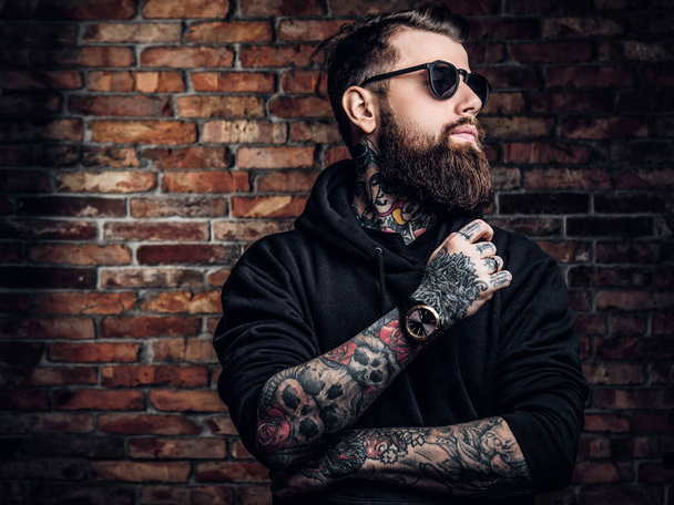 Ένα κομψό τατουάζ ο τύπος σε ένα μαύρο φούτερ με κουκούλα και γυαλιά ηλίου. Στούντιο φωτογραφία ενάντια στον τοίχο - Φωτογραφία, εικόνα