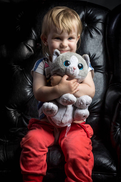 funny cute baby kit girl portrait with different emotions in red pants with soft toy sitting in black leather chair in studio with black isolated background - Φωτογραφία, εικόνα