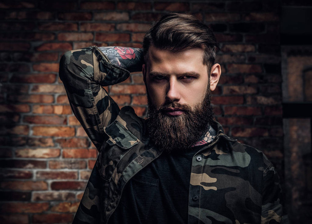 Портрет татуйованого бородатого хлопця у військовій сорочці. Студійне фото проти цегляної стіни
 - Фото, зображення