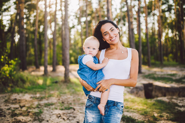 junge schöne modische Mutter mit langen brünetten Haaren hält eine Tochter Blondine mit blauen Augen ein Jahr nach der Geburt in einem Nadelwald in einem Sommerpark. - Foto, Bild