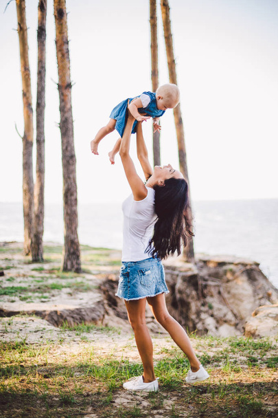 Mothers Day. A beautiful young Caucasian woman mommy holds on her hands, throws up lifts on her hands up daughter in the summer in the forest. Mom and child in nature in the forest park - Foto, Imagen