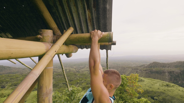 Сильный человек тренировки тянуть вверх упражнения на деревянном баре на холме и высокогорных покрытых тропических лесов. Спортивный человек подъезжает к горизонтальной перекладине на тропическом ландшафте. Спортивный зал на открытом воздухе
. - Кадры, видео
