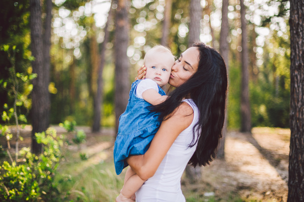 young beautiful fashionable mother with long brunette hair holding a daughter's blonde with blue eyes one year of birth in a coniferous forest in a summer park. Wearing jeans clothes - Photo, Image