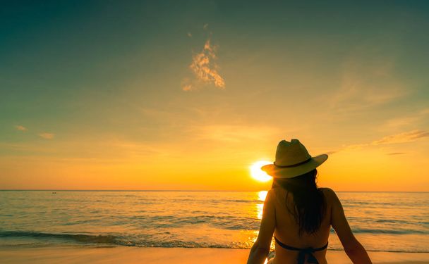 Back view of Asian woman wear bikini and straw hat sit on sand beach relax and enjoy holiday at tropical paradise beach. Woman watch beautiful sunset. Summer vacation. Travel alone. Summer vibes. - Photo, image