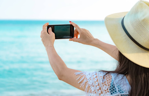 Азиатская женщина хипстер носить шляпу использовать смартфон, фотографируя красивое море. Летние каникулы на пляже тропического рая. Счастливая девушка-хипстер путешествует в отпуске. Женщина наслаждается и расслабляется. Летние вибрации
.  - Фото, изображение
