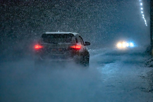 Το αυτοκίνητο πηγαίνει στο δρόμο σε μια απρόσμενη άνοιξη χιονόπτωση το βράδυ. - Φωτογραφία, εικόνα