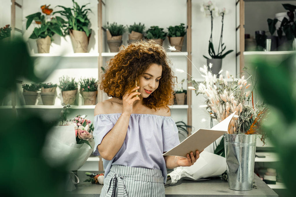 Curly worker of floral shop looking into notebook and calling client - Photo, image
