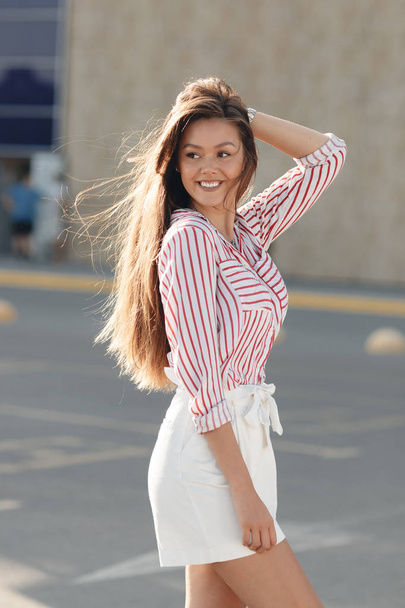 Fashion portrait of a sensual woman. External photo of an attractive brunette model in a white skirt and a striped blouse. Gorgeous portrait of a beautiful sexy woman outdoors in the summer on the city street. Natural beauty and lifestyle concept - Фото, изображение
