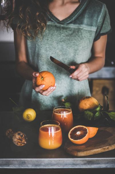 Young female making freshly squeezed blood orange juice or smoothie near concrete kitchen counter. Healthy lifestyle, vegan, vegetarian, alkaline diet concept - 写真・画像
