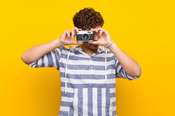 Blonde man over yellow wall holding a camera - 写真・画像