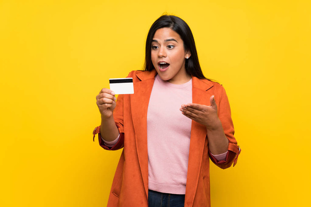 Νεαρό Κολομβιανό κορίτσι πάνω από κίτρινο τοίχο κρατώντας μια πιστωτική κάρτα και έκπληκτος - Φωτογραφία, εικόνα