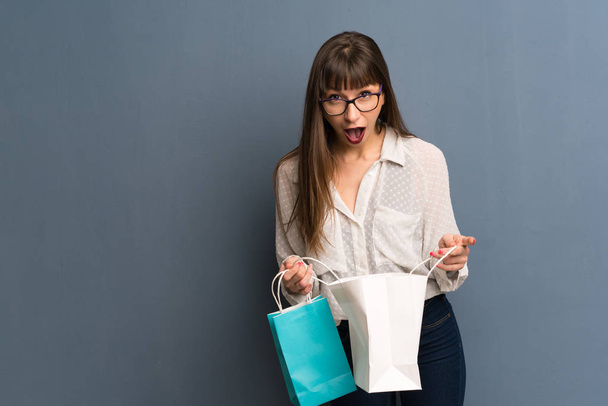 Woman with glasses over blue wall surprised while holding a lot of shopping bags - Фото, изображение