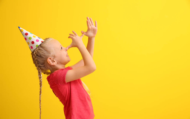 Funny little girl in party hat grimacing on color background. April fools' day celebration - Photo, Image
