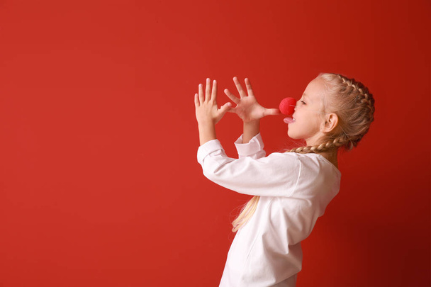 Funny little girl with clown nose on color background. April fools' day celebration - 写真・画像