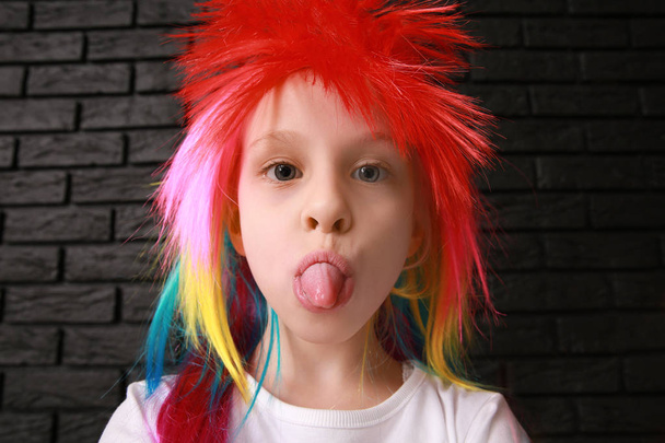 Funny little girl in bright wig showing tongue on dark background. April fools' day celebration - Photo, Image