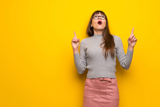 Woman with glasses over yellow wall surprised and pointing up - Photo, Image