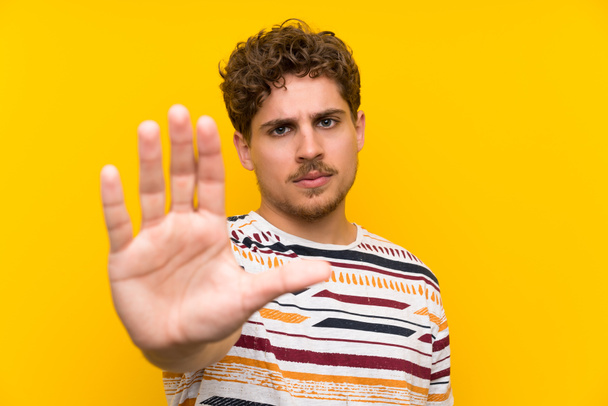 Hombre rubio sobre pared amarilla haciendo gesto de parada con su mano
 - Foto, Imagen
