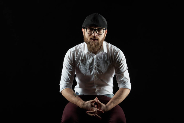 Portrait of a young man in glasses with a beard and a mustache in a black cap and a white shirt, A dark studio background  - Valokuva, kuva