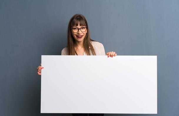 Vrouw met bril over blauwe muur houdt een lege bordje voor een concept invoegen - Foto, afbeelding