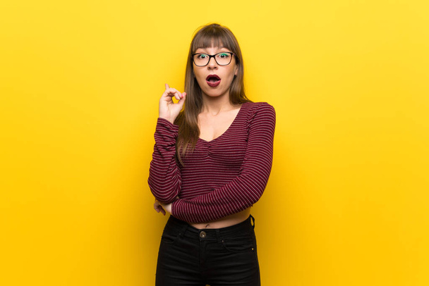 Mujer con gafas sobre pared amarilla pensando en una idea apuntando con el dedo hacia arriba
 - Foto, imagen