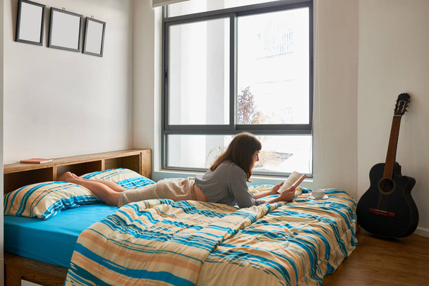 Giovane donna sdraiata sul letto con tazza di caffè e libro interessante
 - Foto, immagini