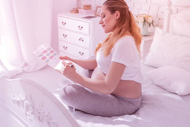 Μακρυμάλλης έγκυος γυναίκα σε ένα λευκό t-shirt σήμανση ημερομηνίες στο ημερολόγιο - Φωτογραφία, εικόνα