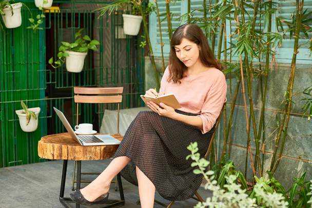 Hymyilevä luova nuori nainen istuu tuolilla takapihallaan ja kirjoittaa runoja päiväkirjaan
  - Valokuva, kuva