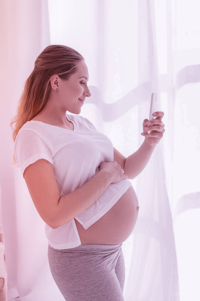 junge strahlende langhaarige schwangere Frau in weißem T-Shirt macht Selfie - Foto, Bild