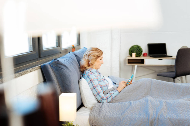 Concentrated adult woman in bright blue pajama leaning on bed - Photo, image