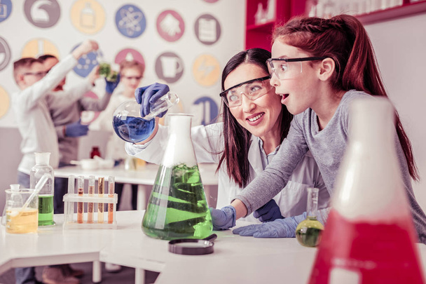 Beaming long-haired teacher mixing liquids in big bulb - Fotó, kép