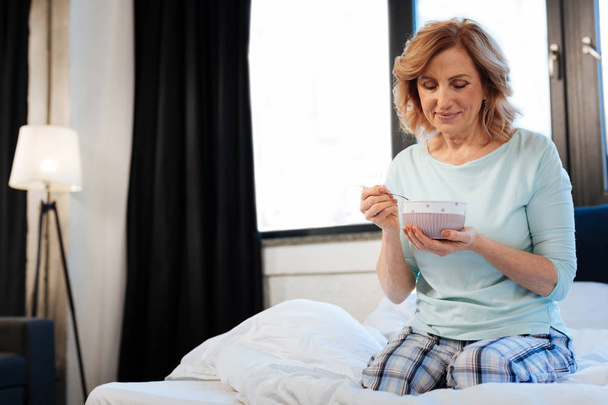 Pleasant short-haired mature lady sitting in bed with bowl of light breakfast - Fotoğraf, Görsel