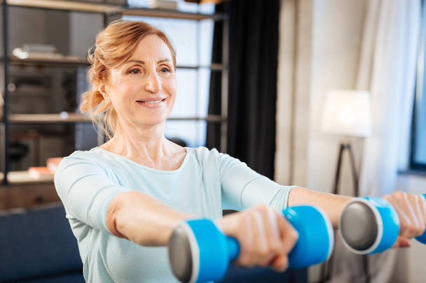Positive adult lady raising heavy dumbbells while having training at home - Foto, afbeelding