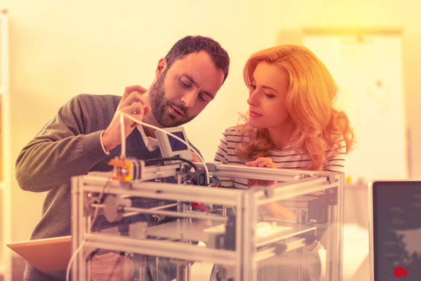 Serious man and woman tinkering with the mechanism of a brand-new 3D printer. - Фото, изображение