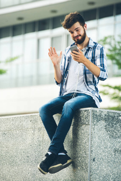 Wesoły, młody człowiek siedzi na zewnątrz i wideorozmów w telefonie komórkowym - Zdjęcie, obraz