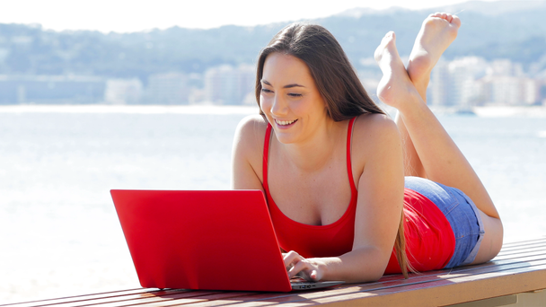 Boldog lány piros használ egy laptop, feküdt egy padon, a partja - Felvétel, videó