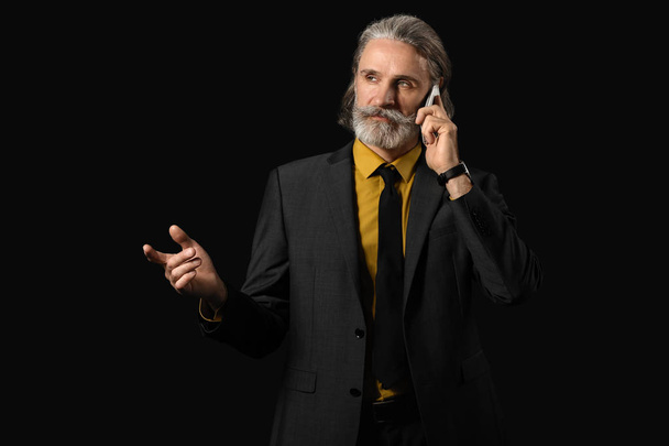 Handsome mature businessman talking by phone on dark background - Photo, Image