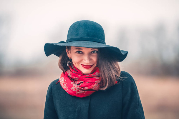 La joven camina en la primavera en el campo vacío con un abrigo negro y un sombrero. Lápiz labial rojo en los labios y una sonrisa. Moda y estilo
 - Foto, imagen