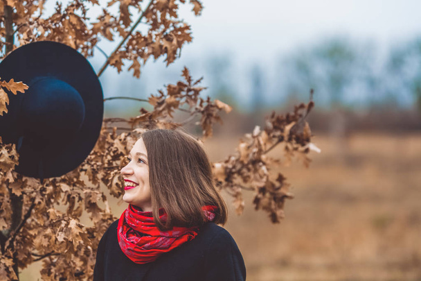 Nuori tyttö kävelee puistossa syksyllä mustassa takissa, hattu roikkuu puussa lähellä. Punainen huulipuna. Muoti ja tyyli
. - Valokuva, kuva